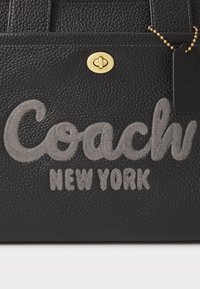 Sac en cuir noir texturé avec logo "Coach New York" brodé gris, fermoir tourniquet doré et étiquette en cuir noir sur chaîne dorée.