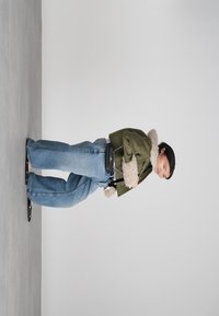 Naistepäev rohelises jopes, millel on karvane äär, laiad sinised teksad ja mustad sandaalid, hoides väikest musta käekotti, seismas halli ühtlase tausta ees.