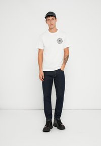 Tricou alb cu un grafic circular negru, blugi de culoare albastră închisă și pantofi negri cu șireturi. Modelul mai poartă o șapcă închisă la culoare.