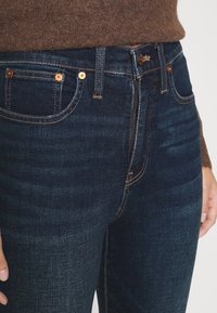 Jeans de mezclilla azul oscuro con remaches y costuras de cobre, combinados con un suéter marrón, centrándose en la cremallera y los bolsillos frontales.