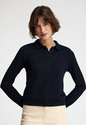 Kobieta o krótkich brązowych włosach, ubrana w czarną, prążkowaną koszulkę polo z długim rękawem i beżowe spodnie, stojąca z rękami na biodrach na jednolitym tle.