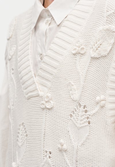 Gilet blanc en maille avec un col en V, orné de broderies florales, de côtes texturées et de détails en relief. Porté par-dessus une chemise de couleur claire.