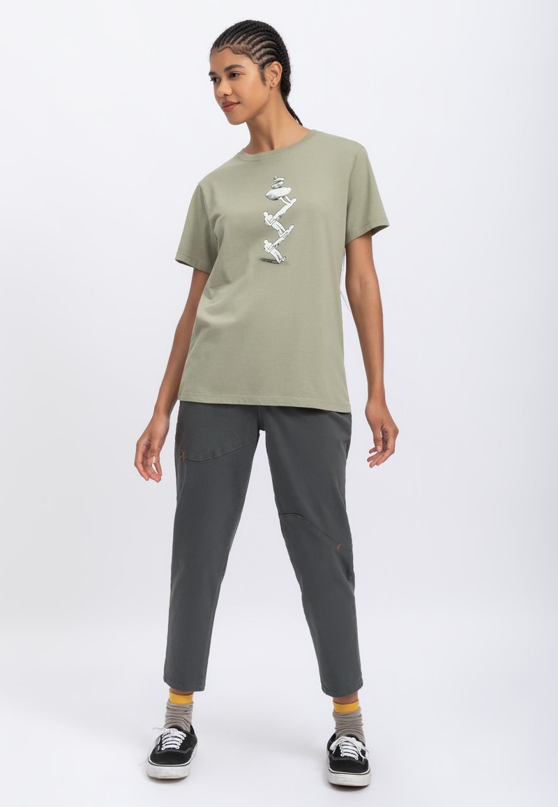 Olijfgroen katoenen t-shirt met een grafisch ontwerp aan de voorkant; gecombineerd met donkergrijze broek met zakken en gestikte details. Casual schoeisel inbegrepen.