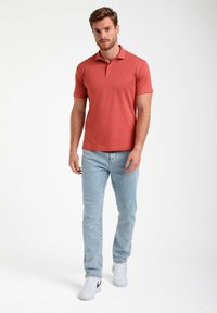Rød polo-shirt med korte ærmer, parret med lyseblå jeans og hvide sportsko, stående på en ensfarvet baggrund.