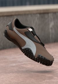 Sneaker Puma maro și negru cu logo alb și talpă texturată, ridicat pe un fundal din beton.