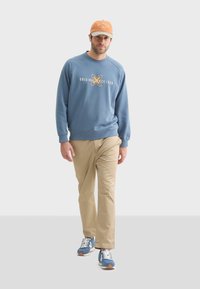 Sudadera azul con el texto blanco "ORIGINAL SINCE 1939", pantalones caqui y zapatillas azules con cordones blancos. Gorro beige con acentos de logo.