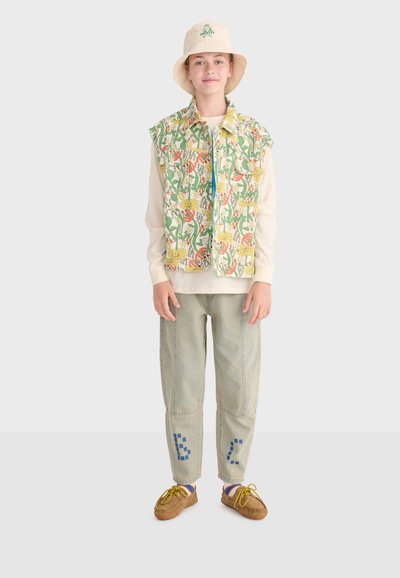 Jeune personne portant un chapeau bob beige, une veste sans manches à motif floral, une chemise à manches longues beige clair, un pantalon gris avec des patchs bleus et des chaussures marron.