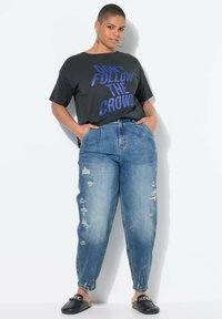 Camiseta negra con texto azul que dice "NO SIGAS A LA MULTITUD", combinada con pantalones jogger de mezclilla azul claro desgastados y zapatos negros sin cordones.