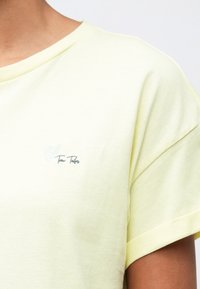 Κοντινό πλάνο ενός ατόμου που φοράει ένα ανοιχτό κίτρινο κοντομάνικο πουκάμισο με ένα μικρό ανοιχτό μπλε και γκρι λογότυπο στο στήθος.