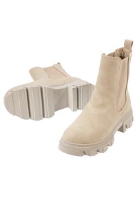 Bottes Chelsea en daim beige avec semelles en caoutchouc épaisses et panneaux latéraux élastiques, une botte debout et l'autre montrant la semelle sur fond blanc.