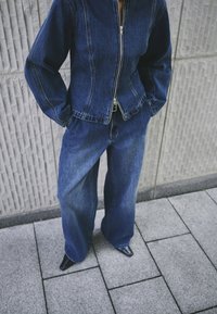 Traper jakna sa patentnim zatvaračem, tamnoplava, široke traperice u odgovarajućem denimu. Glatka tekstura i vidljive šavove, uparen sa špicastim crnim cipelama.