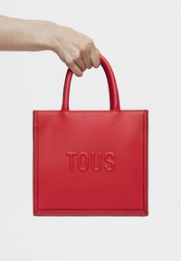 Bolso tote de cuero rojo con el logo "TOUS" en relieve, forma rectangular estructurada y dos asas redondeadas. Textura suave con costuras en contraste.