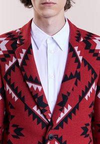 Blazer en laine rouge avec un motif géométrique noir et blanc audacieux, doté d'un col cranté et d'une fermeture à un bouton, porté sur une chemise blanche.