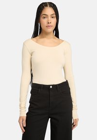 Beige lange mouw bodysuit met een off-the-shoulder halslijn, gecombineerd met high-waisted zwarte jeans. Gladde textuur, strakke pasvorm.