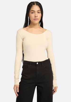 Beige lange mouw bodysuit met een off-the-shoulder halslijn, gecombineerd met high-waisted zwarte jeans. Gladde textuur, strakke pasvorm.