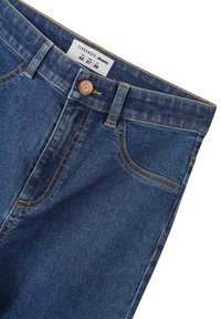 Jeans in denim blu scuro con cuciture dorate a contrasto, dotati di un bottone di rame, cinque tasche e un'etichetta che mostra la taglia e il marchio.