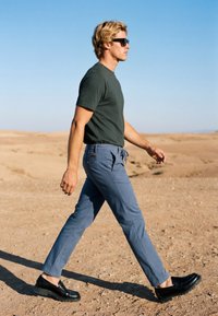 Homme blond portant des lunettes de soleil, un t-shirt vert foncé, un pantalon bleu et des chaussures noires marchant sur un terrain sec et sablonneux sous un ciel bleu clair.