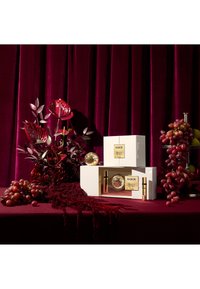 Tre scatole bianche con accenti dorati espongono prodotti per la cura della pelle, circondate da fiori rossi, uva nera e uno sfondo di velluto burgundy.