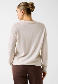 Jasnobeżowy sweter o luźnym kroju, z prążkowanym dekoltem i mankietami, noszony na brązowych dżinsach. Miękka faktura, minimalistyczny design, bezszwowe plecy.