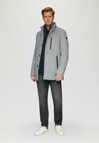 Grijze wollen trenchcoat met hoge kraag, voorzien van contrasterende zwarte ritssluitingen en knopen, gecombineerd met donkere jeans en witte sneakers.