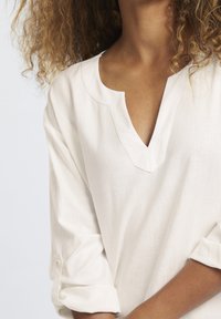 Biała lniana bluzka z dekoltem w kształcie litery V i podwiniętymi rękawami. Materiał wydaje się miękki o lekko fakturowanej powierzchni. Prosty i minimalistyczny design.