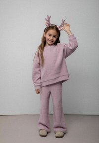 Młoda dziewczyna w miękkim różowym dzianinowym swetrze i spodniach, z błyszczącym opaską z rogami renifera, uśmiechająca się i stojąca na neutralnej podłodze.