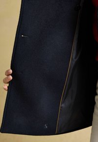 Tumesinine villane mantel, millel on sujuv disain, sisemine vooder kontrastse värviga, peened õmblused ja väike logo detail.