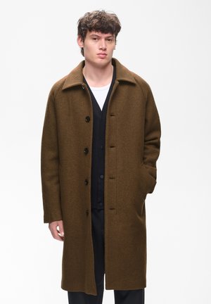Jeune homme portant un long manteau marron boutonné par-dessus une tenue noire, une main dans la poche, debout devant un fond blanc uni.