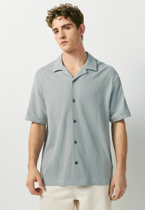 Junger Mann trägt ein hellgraues, kurzärmeliges Hemd mit Knöpfen und einem strukturierten Chevron-Muster sowie beige Hosen, steht vor einem schlichten Hintergrund.