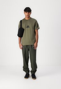 Olivenfarget treningst-skjorte med svart logo, kombinert med matchende smale joggebukser. Bært med svarte joggesko og en liten svart skulderveske.