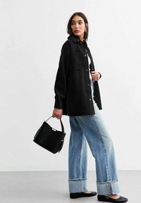 Camicia overshirt nera con due tasche sul petto, abbinata a jeans a gamba larga di colore blu chiaro. Una borsa nera e scarpe basse completano l'outfit.