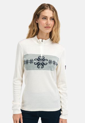 Kvinne med blondt hår iført en hvit langermet genser med grått og marineblått snøfnuggmønster og frontglidelås, stående mot en ensfarget bakgrunn.