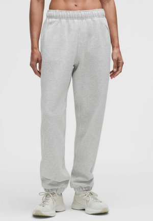 Osoba nosząca jasno szare spodnie dresowe z elastycznym pasem i białe sportowe trampki, stojąca na jednolitym jasnym tle.