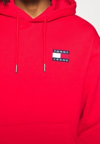 Felpa rossa con cappuccio, dotata di una tasca frontale a forma di canguro e di un'etichetta rettangolare nera con la scritta "TOMMY" e "JEANS". Texture liscia e cordini regolabili.