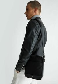 Borsa a tracolla nera realizzata in materiale resistente, forma rettangolare, con una tracolla sottile e tasca frontale. Texture liscia, design minimalista.