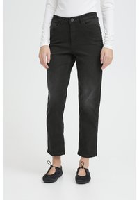 Osoba oblečená v čiernych džínsach s rovnými nohavicami, čiernych baletných balerínkach s očkami a bielom tričku s dlhým rukávom, stojaca na bielom pozadí.