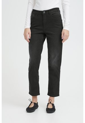 Person iført sorte jeans med lige ben, sorte ballerinasko med stropper og en hvid bluse med lange ærmer, stående på en hvid baggrund.