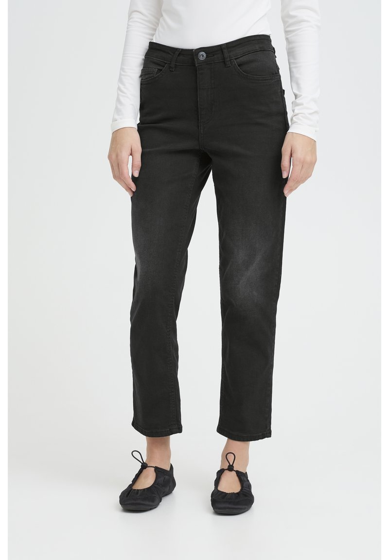 Osoba oblečená v čiernych džínsach s rovnými nohavicami, čiernych baletných balerínkach s očkami a bielom tričku s dlhým rukávom, stojaca na bielom pozadí.