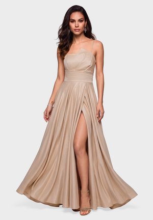 Femme portant une robe de soirée beige sans manches avec une fente haute, debout avec une jambe visible et de longs cheveux ondulés et foncés.