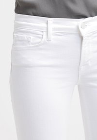 Jeans de mezclilla blanca con corte ajustado. Las características incluyen un cierre de botón, bolsillos frontales y una tela suave y texturizada.