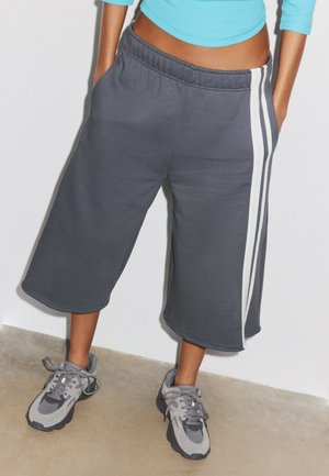Personne portant un short gris foncé arrivant aux genoux avec des bandes blanches sur les côtés, un haut bleu court à manches longues, et des baskets de sport grises, debout à l'intérieur.