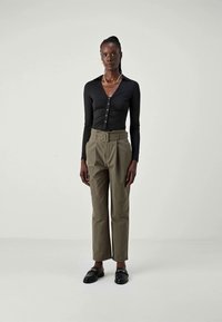 Black long-sleeve top with buttons, paired with olive-green high-waisted trousers featuring a belt, and black loafers on a neutral background.