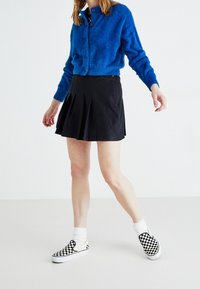 Niebieski puszysty kardigan, czarna plisowana spódnica oraz czarno-białe sznurowane buty w kratę z białymi skarpetkami. Modelka idzie na jednolitym tle.