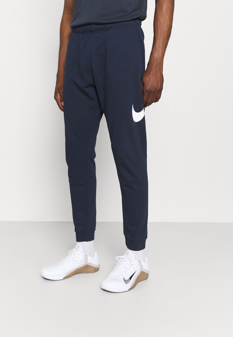 Pantalons de jogging bleu marine avec un logo Nike blanc sur la cuisse gauche, taille élastique et poignets. Associés à des chaussures de sport blanches.