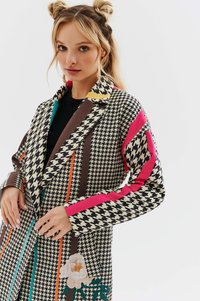 Płaszcz w wzór houndstooth w kolorach czarnym, beżowym i brązowym. Posiada kolorowe akcenty, hafty kwiatowe oraz kołnierz z wycięciem.