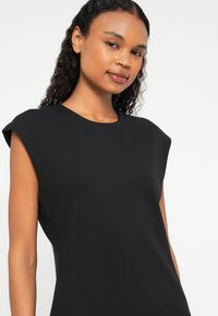 Melna kleita ar augstu kakla izgriezumu un īsām piedurknēm. Mīksts audums ar gludu tekstūru, pieguļoša silueta un vienkāršs dizains.