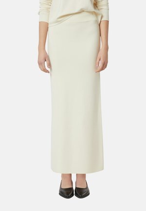 Kobieta ubrana w kremową długą spódnicę, kremowy sweter i czarne szpilki, stojąca na tle gładkiego, białego tła.