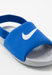 Des sandales bleues à enfiler avec une semelle texturée grise, dotées d'une large sangle bleue et d'un logo Nike blanc. Matériau synthétique doux et bande élastique.