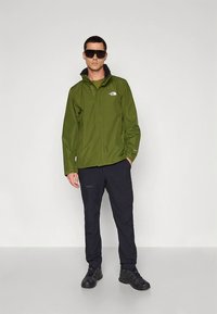 Casaco impermeável verde com gola alta e detalhes pretos, usado sobre calças e calçado pretos. Modelo usa óculos escuros, em posição ereta.