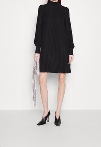 Robe noire à manches longues avec un col montant, fabriquée dans un tissu métallique. Associée à des talons noirs à bouts pointus et une pochette argentée à franges.